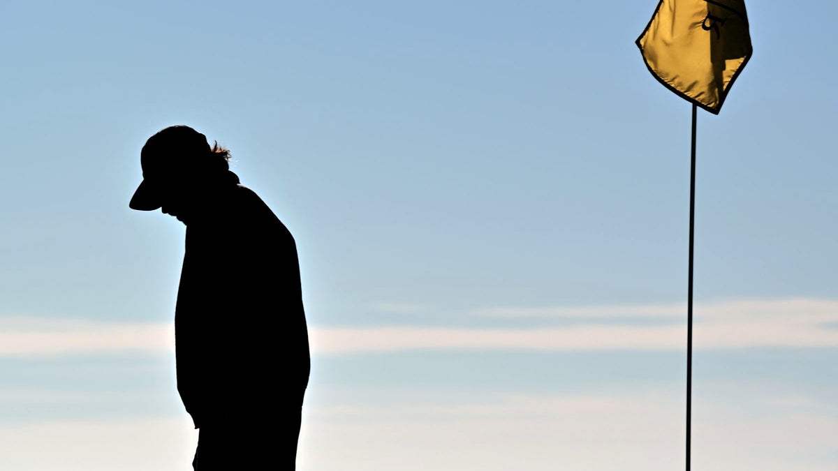 A lone golfer in silhouette at dusk, head bowed, standing beside the flagstick on a green