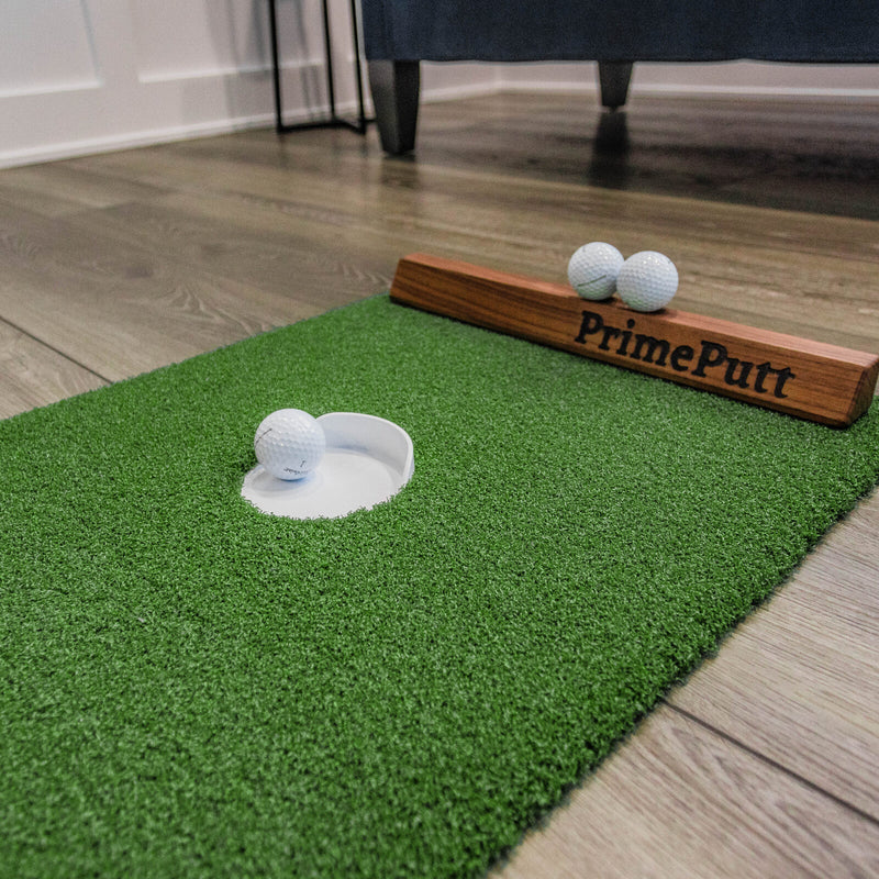 Golf ball resting in the cup of the PrimePutt Compact putting mat beside the walnut PrimePutt backstop