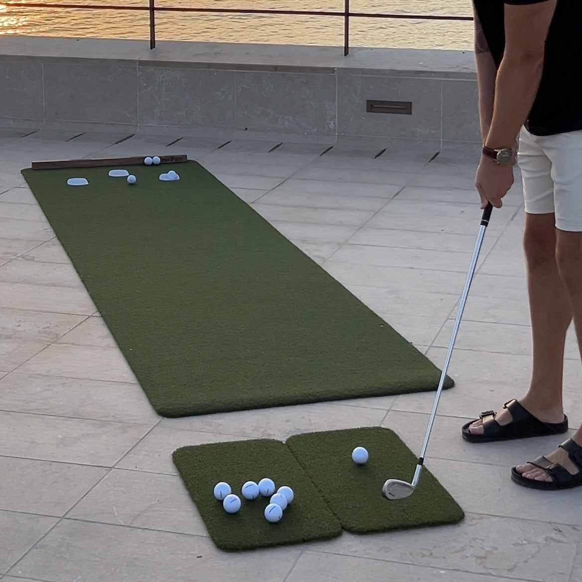 Roll-out putting mat laid flat on a seaside terrace at sunset, with a golfer putting from a hitting mat